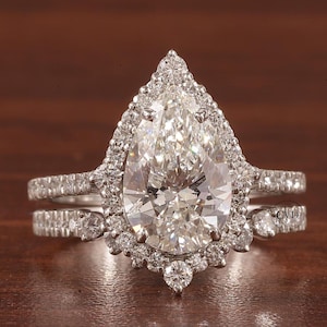 May include: A pear-shaped diamond engagement ring with a halo setting and a matching wedding band. The ring is set in white gold and features a large, sparkling diamond.