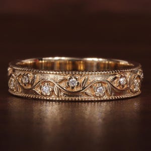 May include: A gold band ring with a floral design. The ring features small, sparkling diamonds set within an intricate pattern of leaves and flowers. The ring is approximately 0.25 inches wide.