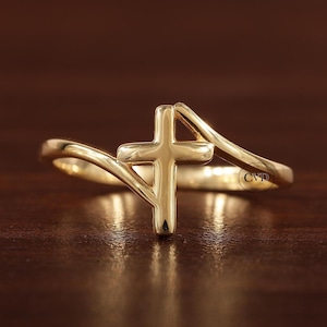 May include: A gold-toned ring featuring a simple cross design. The band of the ring is twisted and the cross is positioned centrally on the band.