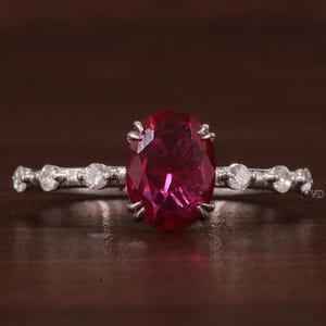 May include: A silver ring featuring a central oval-cut deep red gemstone held by six prongs. The band is accented with three small round clear stones on each side of the center setting, creating a symmetrical design with a polished metallic finish.