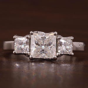 May include: A three-stone ring featuring a large central princess-cut clear gemstone flanked by two smaller princess-cut clear gemstones. The stones are set in a polished silver-toned metal band with a small CVD inscription visible on the side.