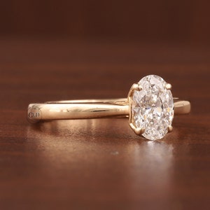 May include: A gold engagement ring featuring an oval-cut diamond. The diamond is held in place by four prongs on a slender, polished gold band. The ring is presented on a dark brown surface.