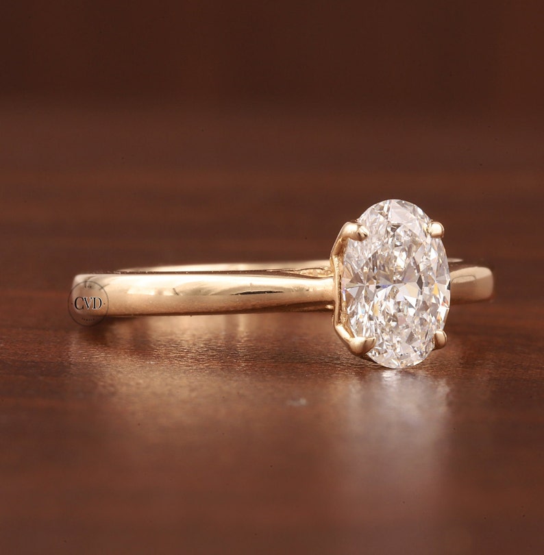 May include: A gold engagement ring featuring an oval-cut diamond. The diamond is held in place by four prongs on a slender, polished gold band. The ring is presented on a dark brown surface.