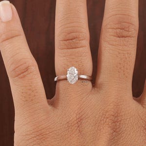 May include: A silver ring featuring an oval-cut diamond. The diamond is set in a simple band. The ring is displayed on a finger, against a brown backdrop.