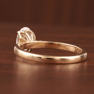 May include: A gold engagement ring with a pear-shaped diamond. The ring has a delicate band and intricate detailing around the diamond setting. The ring is shown on a brown surface.