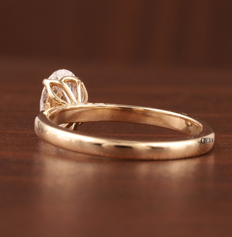 May include: A gold engagement ring with a pear-shaped diamond. The ring has a delicate band and intricate detailing around the diamond setting. The ring is shown on a brown surface.