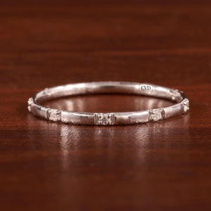 Pode incluir: Um anel de banda de prata com uma fileira de pequenas pedras transparentes engastadas em uma banda texturizada.