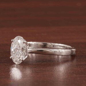 May include: A silver ring featuring an oval-cut diamond. The diamond is clear and reflects light. The ring has a four-prong setting and a polished silver band. The background is a dark brown, wooden surface.