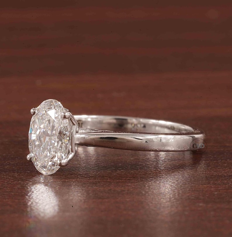May include: A silver ring featuring an oval-cut diamond. The diamond is clear and reflects light. The ring has a four-prong setting and a polished silver band. The background is a dark brown, wooden surface.