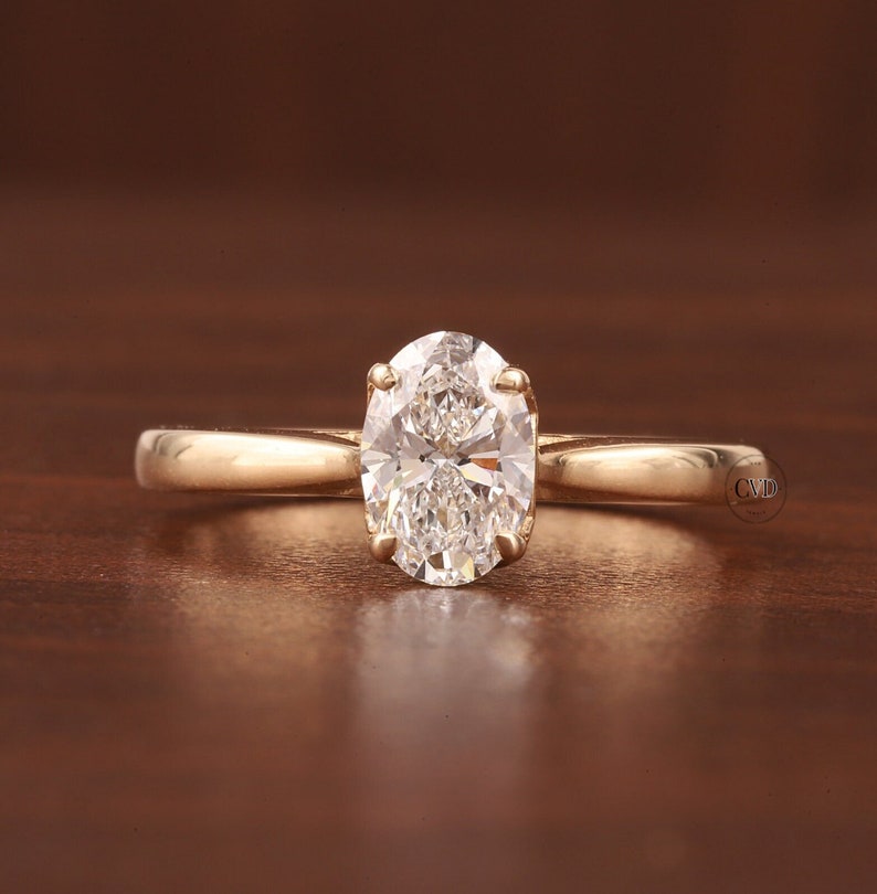 May include: A gold engagement ring featuring an oval-cut diamond. The diamond is held in place by four prongs. The band is plain gold. The ring is presented on a wooden surface. The ring's design is simple and elegant.
