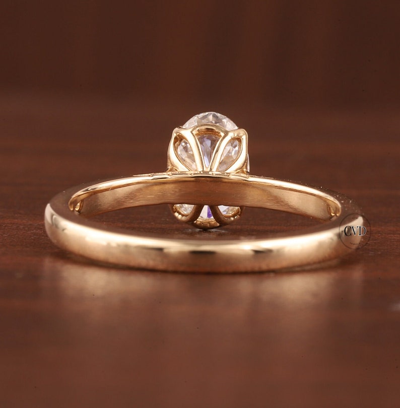 May include: A gold engagement ring with a round, clear gemstone. The ring has a delicate, floral-inspired setting that holds the central stone. The band is smooth and polished, reflecting the light.