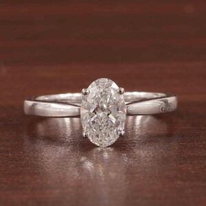 May include: A solitaire engagement ring featuring an oval-cut diamond. The diamond is held in place by four prongs on a polished, silver-coloured band. The ring is shown on a brown surface.