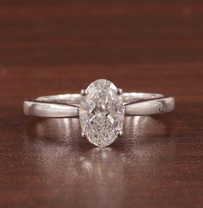 May include: A solitaire engagement ring featuring an oval-cut diamond. The diamond is held in place by four prongs on a polished, silver-coloured band. The ring is shown on a brown surface.