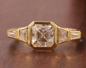 Può includere: Un anello d'oro con una grande pietra preziosa quadrata trasparente circondata da pietre preziose trasparenti più piccole. L'anello ha un design semplice ed elegante.