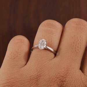 May include: A silver ring featuring a clear, oval gemstone. The ring is set on a slender band and is displayed on a finger. The gemstone is the central element, reflecting light.