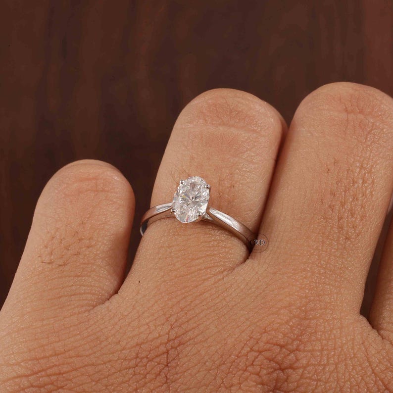 May include: A silver ring featuring a clear, oval gemstone. The ring is set on a slender band and is displayed on a finger. The gemstone is the central element, reflecting light.