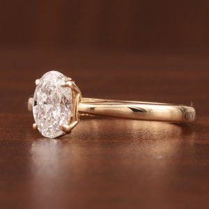 May include: A gold engagement ring featuring an oval-cut diamond. The diamond is held in a four-prong setting. The ring rests on a brown surface. The ring is marked with the letters "CVD" on the side.