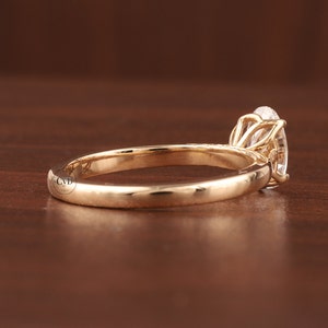May include: A gold engagement ring with an oval-cut diamond. The ring has a slender band and a four-prong setting, holding the gemstone. The ring is shown on a wooden surface, highlighting its elegant design. The diamond is a prominent feature.