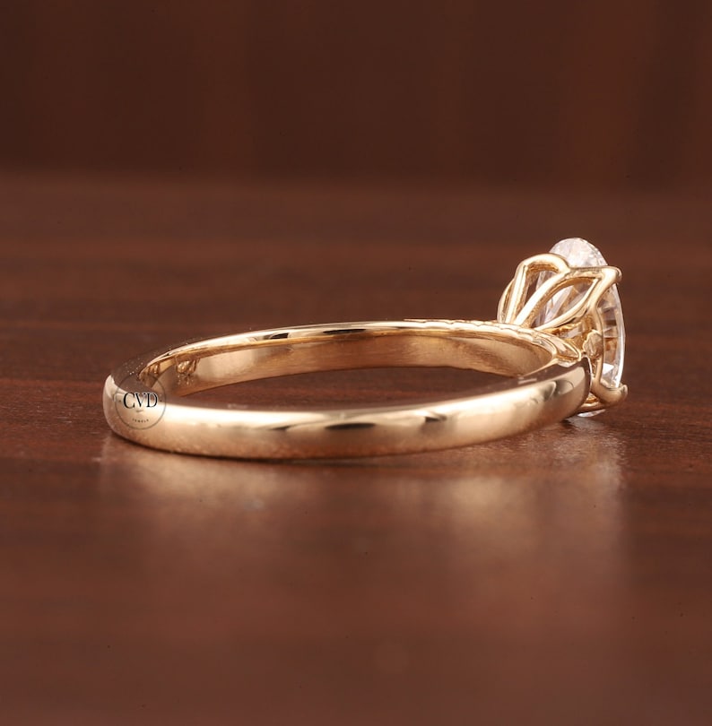 May include: A gold engagement ring with an oval-cut diamond. The ring has a slender band and a four-prong setting, holding the gemstone. The ring is shown on a wooden surface, highlighting its elegant design. The diamond is a prominent feature.