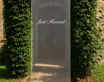 Pode incluir: Uma faixa branca transparente com as palavras "Just Married" em escrita cursiva. A faixa é suspensa por fitas brancas, em um fundo de folhagem verde e um edifício de pedra, tema de casamento.