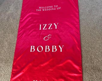Pode incluir: Uma faixa de boas-vindas de casamento vermelha vibrante com texto branco. A faixa diz "WELCOME TO THE WEDDING OF" seguido de "IZZY & BOBBY" e a data "22.08.2026". A faixa é feita de um material brilhante, semelhante ao cetim.