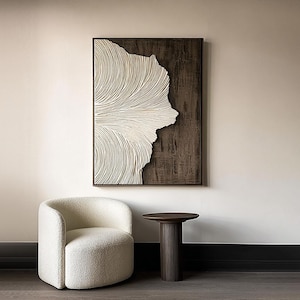 May include: A framed abstract wall art piece with a white, textured, fan-like design against a dark brown background. A white, rounded armchair and a dark brown side table with a ribbed base are in the foreground.