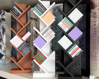 Könnte beinhalten: Drei dekorative Bücherregal-Organizer in Braun, Weiß und Schwarz, jeweils in Form eines stilisierten Baumes. Die Organizer halten Miniaturbücher mit farbigen Buchrücken. Der braune Organizer hat "2025" auf der Basis, der weiße "TBR".