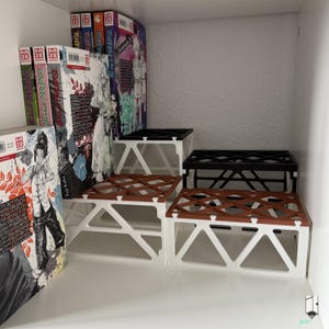 Pode incluir: Organizadores de prateleira de três níveis em branco, castanho e preto, com um design geométrico. Os organizadores estão numa prateleira branca com vários livros de mangá com capas coloridas. Os livros são intitulados "Hell's Paradise".