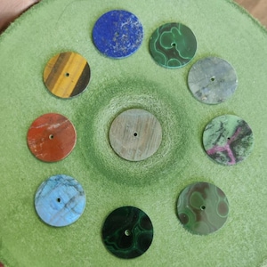 Pode incluir: Uma coleção de dez cabochões de pedras preciosas redondos em várias cores e padrões, incluindo lápis-lazúli azul, malaquite verde e olho de tigre. Cada pedra tem um pequeno furo perfurado no centro. As pedras preciosas estão dispostas sobre uma superfície verde.