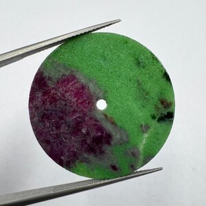 Può includere: Una perla circolare e piatta in pietra preziosa caratterizzata da una base in zoisite verde brillante con una macchia prominente e strutturata di rubino magenta intenso. La pietra presenta un piccolo foro circolare centrale ed è tenuta da pinzette.