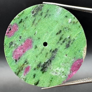 May include: A circular, flat disc of green zoisite stone featuring embedded patches of vibrant pink ruby and dark green to black mineral inclusions. The disc has a small, centered circular hole and is held by metal tweezers.