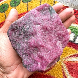May include: A large, raw, unpolished ruby specimen featuring a vibrant magenta-pink crystalline structure with dark grey and black mineral inclusions throughout the rough, irregular surface of the stone, showcasing natural texture and deep color.