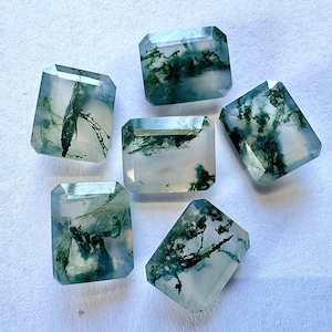 May include: Six rectangular moss agate gemstones with faceted edges are displayed. The translucent stones have a clear base with green, fern-like inclusions. The gemstones are arranged on a white surface, showcasing their unique patterns and clarity.