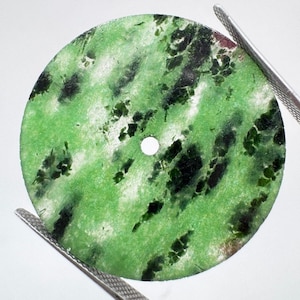 Puede incluir: Un disco circular y plano de piedra verde con un patrón moteado de inclusiones de color verde bosque oscuro y negro. El disco presenta un pequeño orificio circular centrado y está sujeto por pinzas metálicas en los bordes superior e inferior.