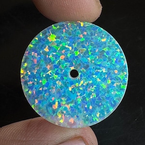 May include: A round, iridescent opal gemstone with a central hole. The opal features a mosaic of blue, green, and yellow colors. The gemstone is held between two fingers against a dark background. This gemstone is suitable for jewelry making.