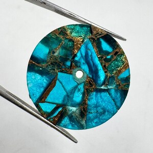 May include: A round, turquoise and bronze-colored stone with a hole in the center. The stone is held by two silver tweezers against a white background. The stone is made of multiple pieces of turquoise and bronze-colored material.