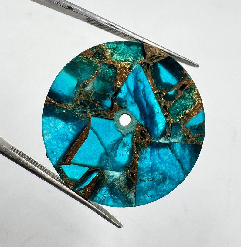 May include: A round, turquoise and bronze-colored stone with a hole in the center. The stone is held by two silver tweezers against a white background. The stone is made of multiple pieces of turquoise and bronze-colored material.
