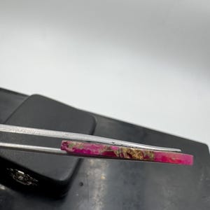 May include: A rectangular gemstone, predominantly pink with gold and brown streaks. Held by silver tweezers, the stone's polished surface is visible against a neutral backdrop. The gemstone's natural patterns are highlighted, suggesting its use in jewellery or decorative items.