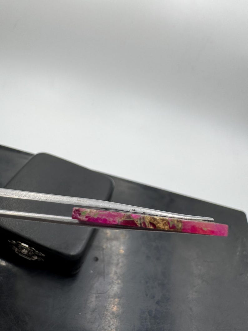May include: A rectangular gemstone, predominantly pink with gold and brown streaks. Held by silver tweezers, the stone's polished surface is visible against a neutral backdrop. The gemstone's natural patterns are highlighted, suggesting its use in jewellery or decorative items.