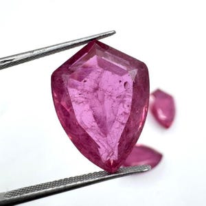 May include: A faceted, shield-shaped, pink gemstone is held by tweezers. The gem has a translucent appearance with internal inclusions. Two additional gems are visible in the background, also pink and shield-shaped. The gems are set against a white background.