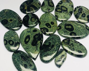 May include: A collection of polished gemstones in various shapes, including oval, teardrop, and oblong. The stones feature a mottled pattern of dark green and black, with a glossy finish. The gemstones are arranged on a white surface.