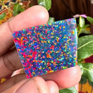 Puede incluir: Una gema rectangular pulida con un mosaico vibrante de colores. La piedra presenta una base azul oscuro salpicada de fragmentos rojos, naranjas, amarillos y verdes, creando un efecto deslumbrante. Los bordes son ligeramente redondeados.