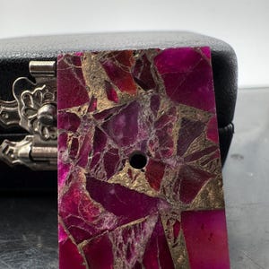 May include: A rectangular, polished stone in a vibrant magenta colour, with a mosaic-like pattern of gold and darker magenta fragments. A small, drilled hole is visible near the top. The stone is likely for jewellery or craft projects.