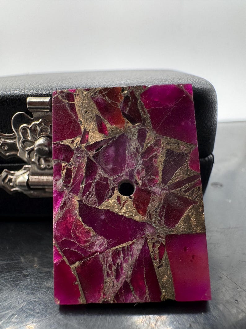 May include: A rectangular, polished stone in a vibrant magenta colour, with a mosaic-like pattern of gold and darker magenta fragments. A small, drilled hole is visible near the top. The stone is likely for jewellery or craft projects.