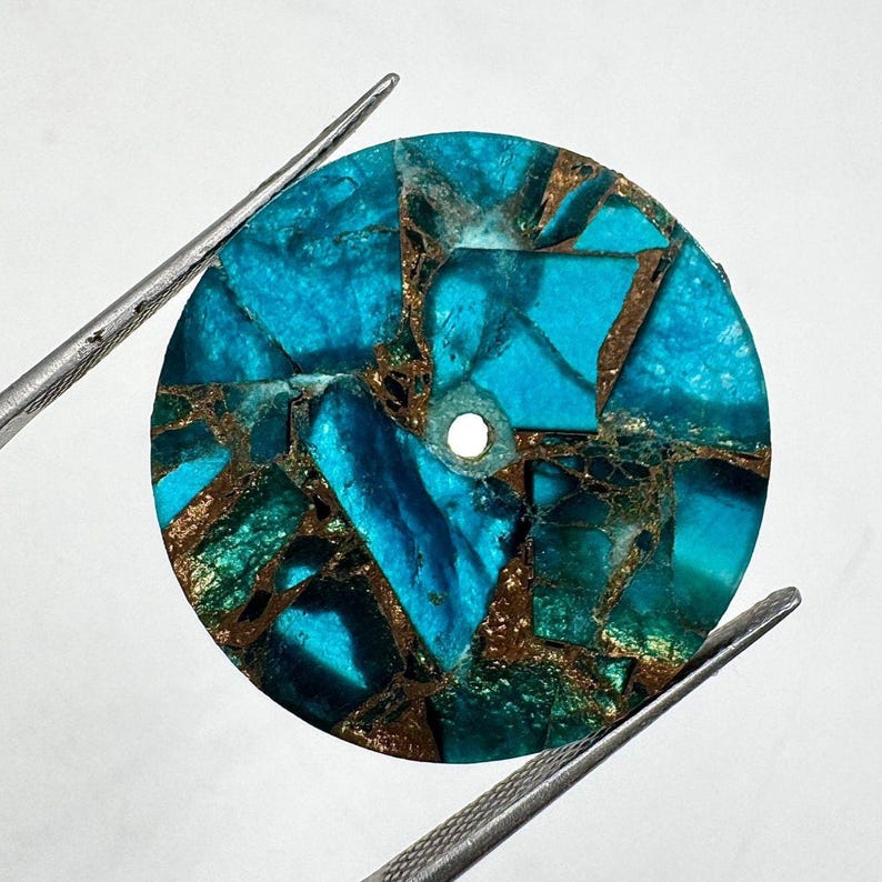 May include: A round, turquoise-colored stone with a mosaic pattern of blue and green fragments, interspersed with bronze-colored veins. A small hole is drilled through the center. The stone is held by two silver tweezers.