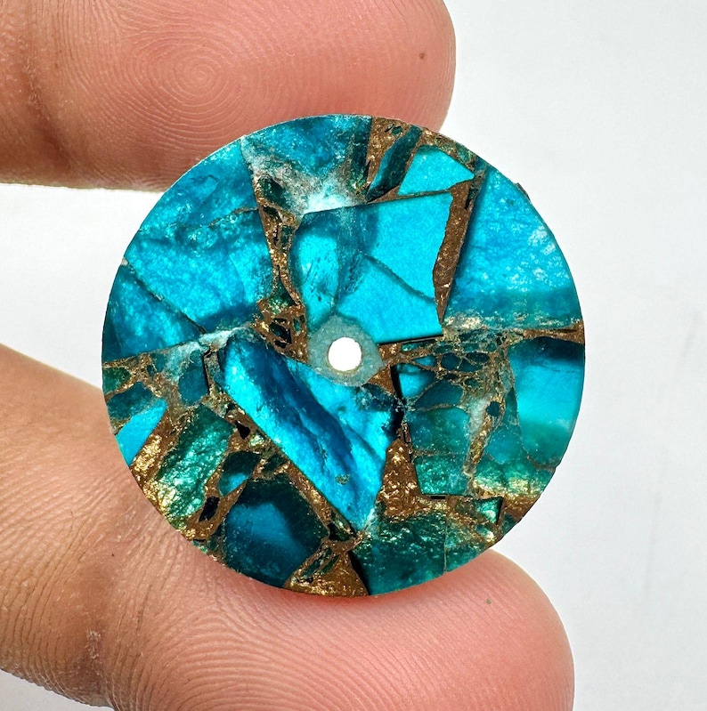 May include: A round, turquoise-colored stone with a mosaic pattern. The stone features a mix of blue and green hues, with gold-colored veins running throughout. A small hole is drilled through the center. The stone is held between two fingers.