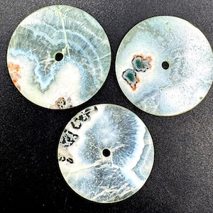 Puede incluir: Tres colgantes de piedra circulares y planos con un orificio central. Cada pieza presenta un patrón natural único en tonos azul pálido, blanco y gris translúcido, con inclusiones que muestran pequeñas manchas de minerales de color naranja, verde y negro.