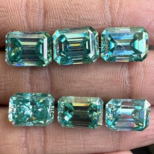 May include: Six emerald-cut gemstones in a light teal hue. The gems are rectangular with faceted edges, reflecting light. The stones are arranged in two rows of three, highlighting their clarity and brilliance.