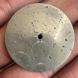 May include: A circular, flat metal disc with a metallic silver-toned finish. The surface features a weathered, speckled texture with dark grey, irregular patches distributed across the face. A single, small, circular hole is centred in the disc.