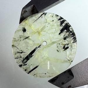 Könnte beinhalten: Ein runder, durchscheinender Edelstein mit schwarzen und gelben Einschlüssen. Der Stein hat ein zentrales Loch und wird von einem Metallwerkzeug gehalten. Die Oberfläche des Edelsteins hat ein rissiges Aussehen.